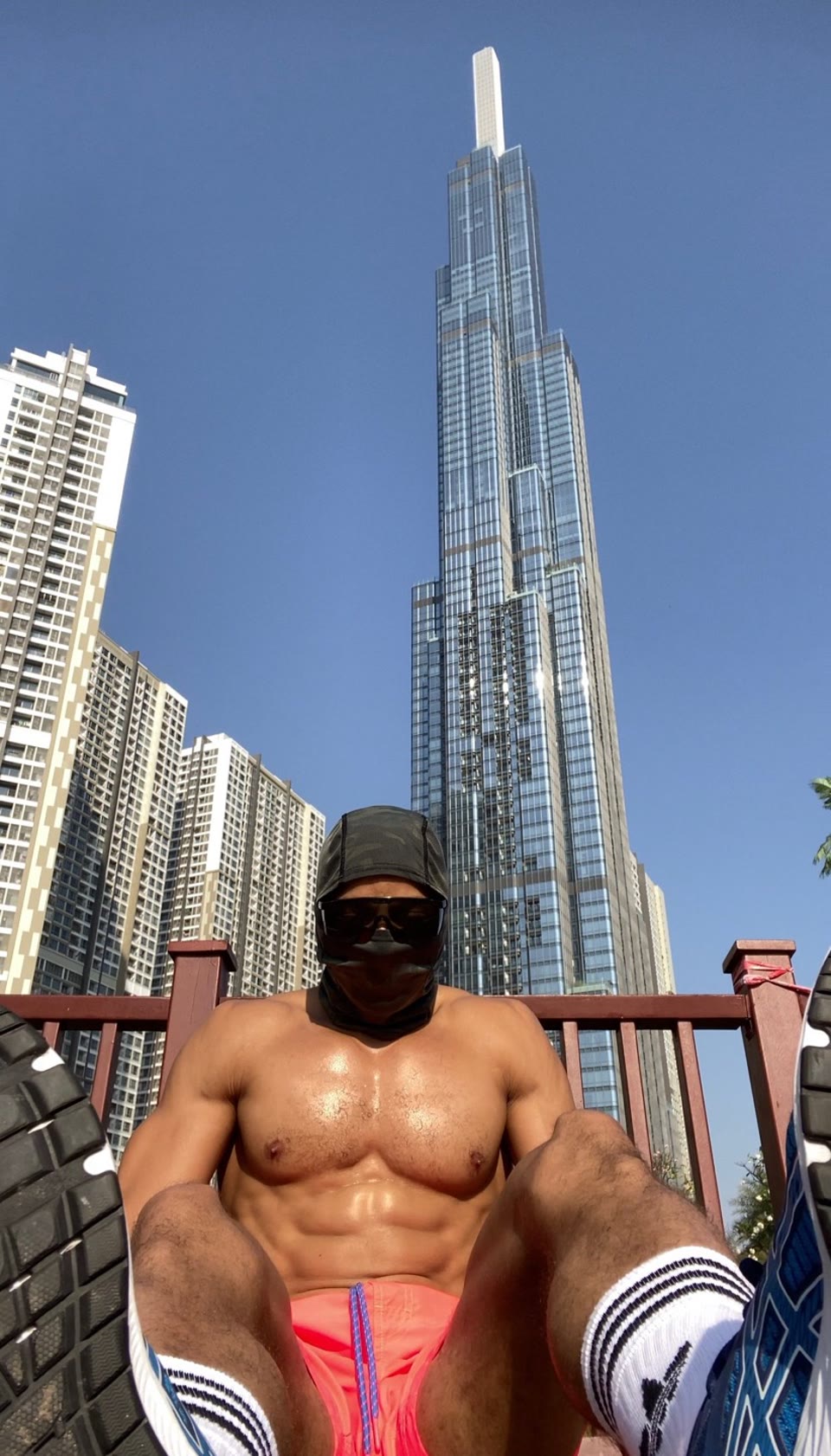 Low-angle outdoor still: masked athlete seated facing the camera, coral shorts and athletic shoes in the foreground, glass skyscrapers and clear blue sky behind a wooden railing.