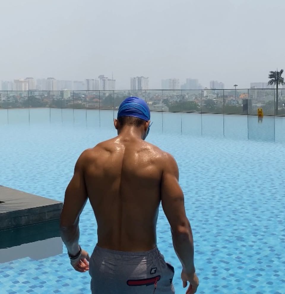 High-angle still from behind: athlete in a blue swim cap standing in a rooftop pool, facing a dense high-rise skyline beyond a glass railing.