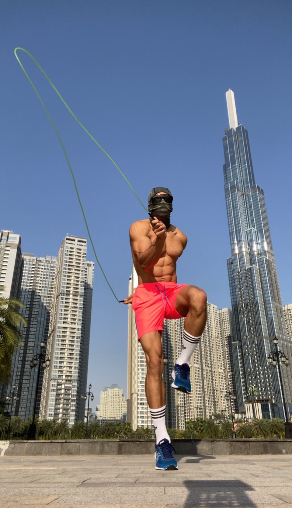 Low-angle outdoor still: athlete mid-jump with a lime-green jump rope on a paved plaza, bright pink shorts, tall glass towers under a clear sky.