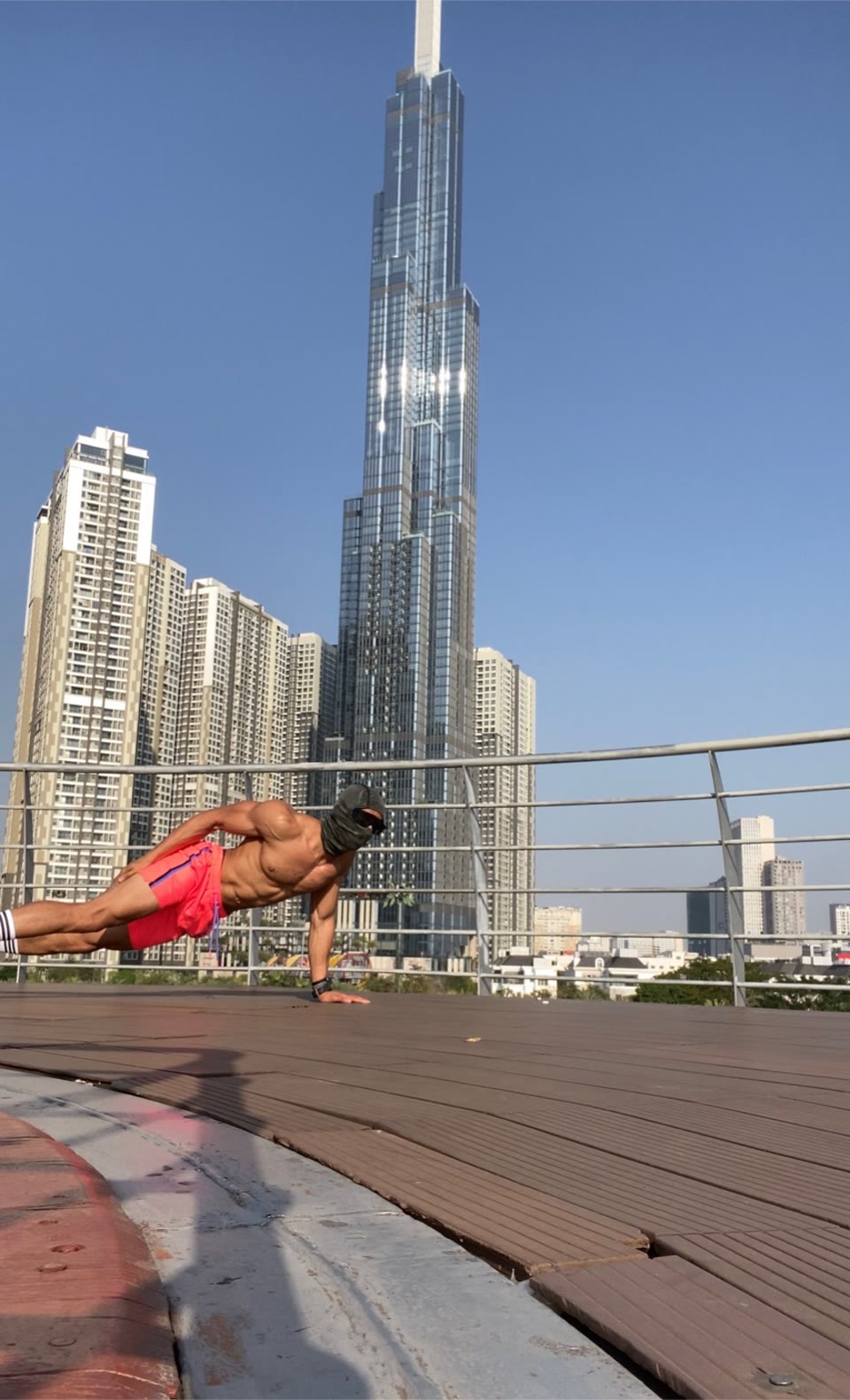 Outdoor deck still: athlete holding a one-arm plank on wooden decking, coral shorts, sunglasses and face covering, tall glass tower and city skyline behind a metal railing.